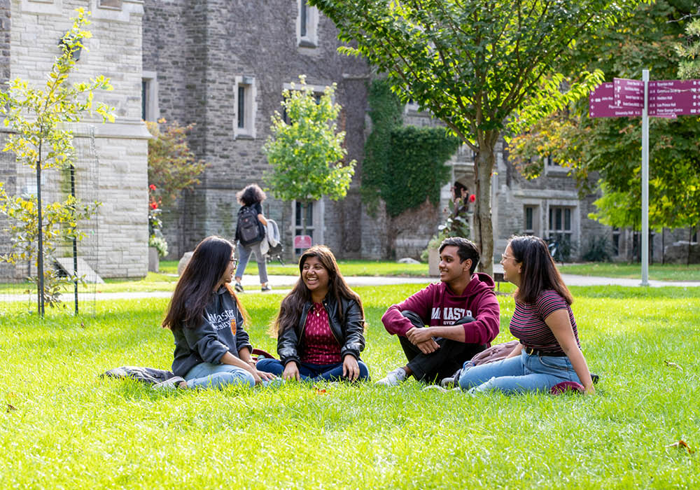 Four young adults sit together on a patch of grass on a summer day at McMaster University in Hamilton, Ontario, Canada.